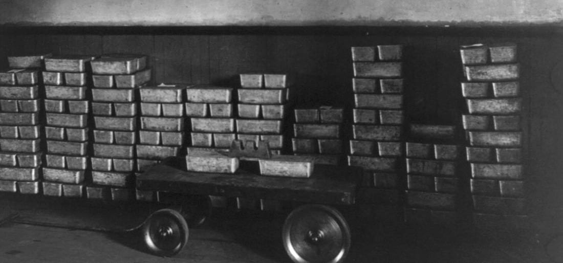 Mehrere Stapel Silberbarren vor einer Wand, davor ein flacher Rollwagen mit weiteren Barren