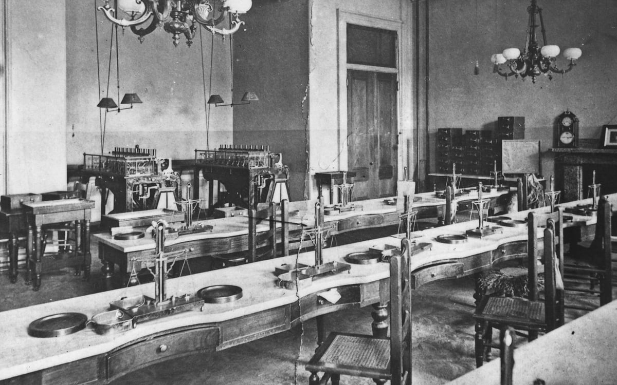 Altes Schwarzweißfoto: ein leerer Saal mit langen Tischen, an jedem Platz eine kleine Waage, hinten Maschinen