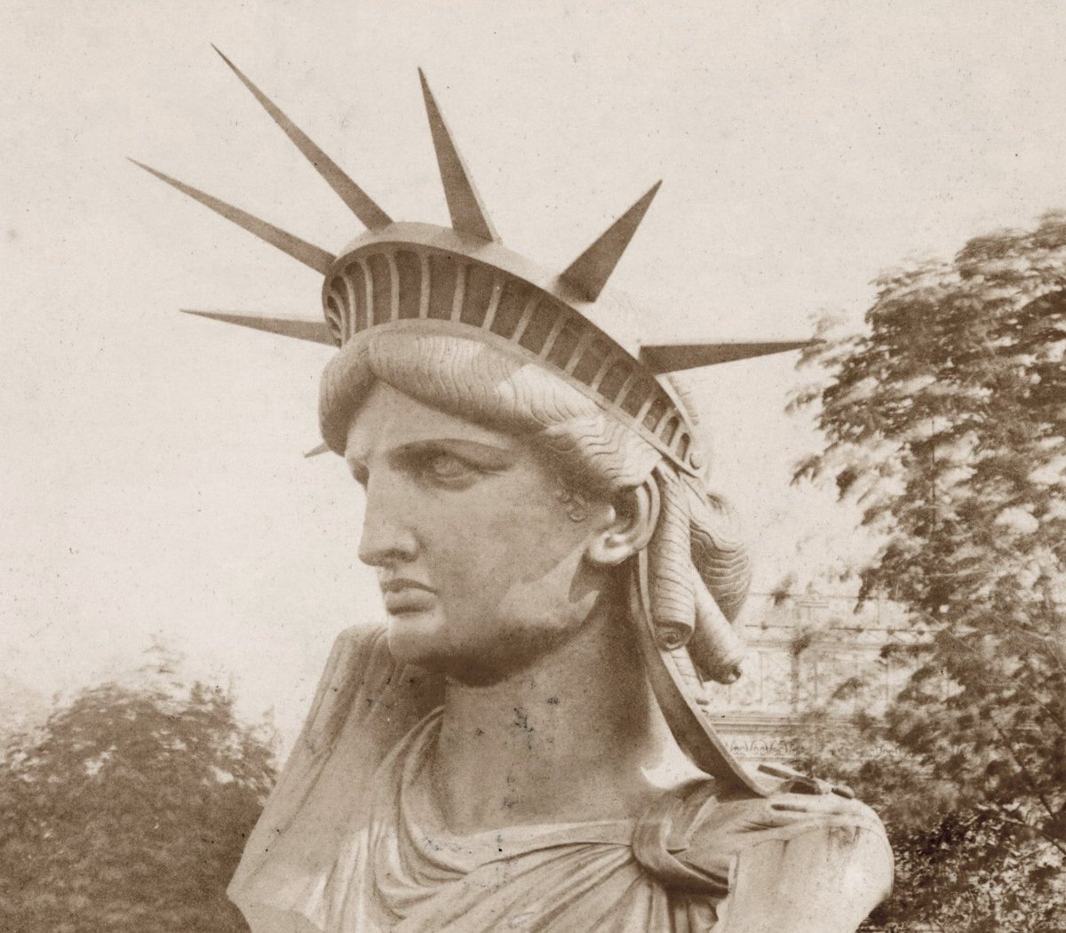 Altes Foto in Sepia: der Kopf der Freiheitsstatue mit Strahlenkrone, im Freien aufgestellt, dahinter Bäume und ein Gebäude