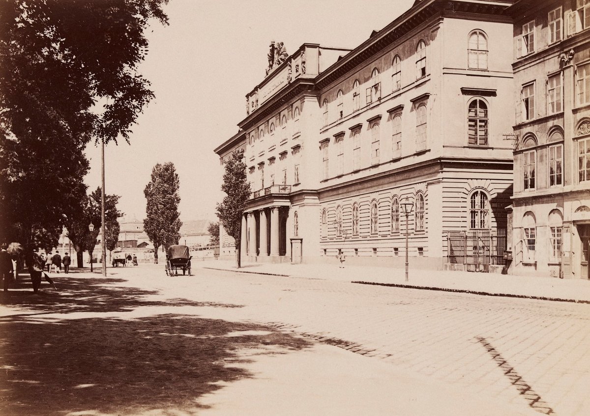 Old photograph: a long neoclassical building with a columned portal on a wide street, trees on the left