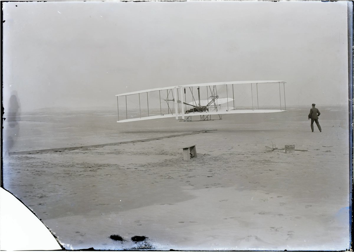 Black‑and‑white photo on a glass plate: the biplane hovers just above the sand, a man lies at the controls on the lower wing, a second man stands on the right. On the left the launching rail leads into the picture; the lower left corner of the glass plate is broken off.