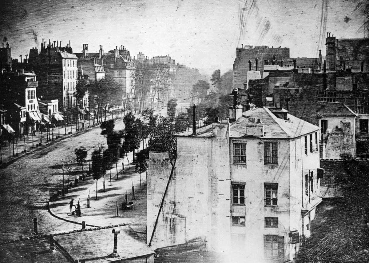Daguerreotypie: Blick von oben auf einen Pariser Boulevard mit Bäumen und hohen Häusern. Die Straße wirkt leer, links unten steht ein Mann beim Schuhputzer.