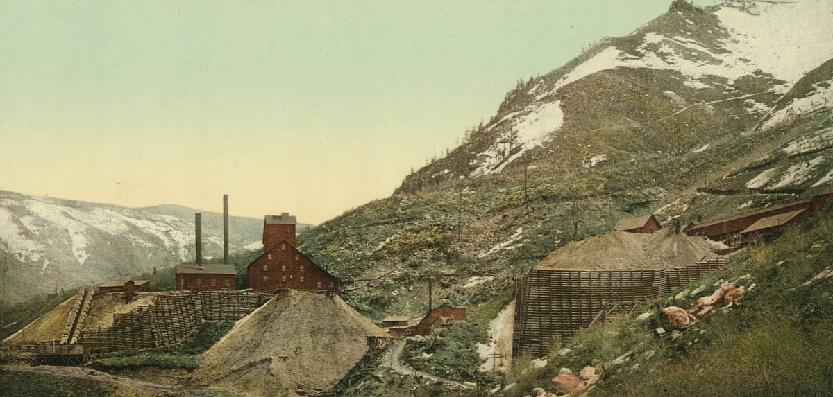 Bergwerk am Hang: rote Fördergebäude mit Schornsteinen, große Halden aus hellem Gestein, dahinter verschneite Berge