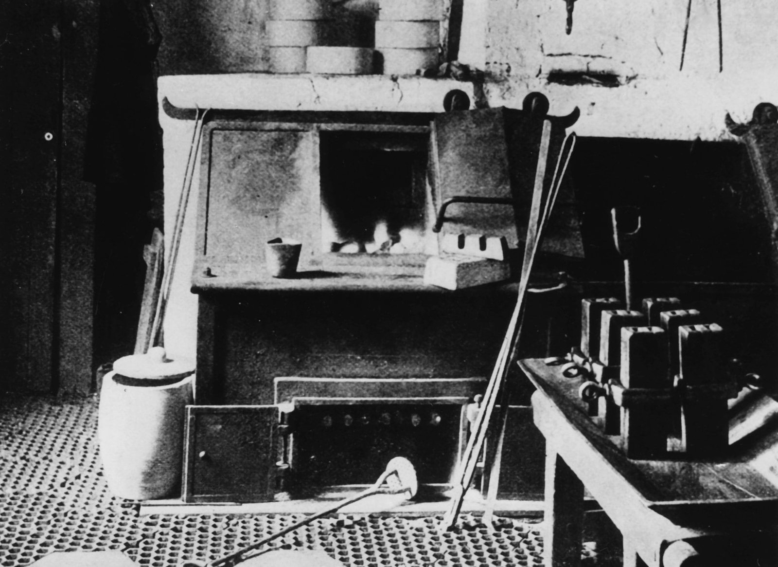 Altes Schwarzweißfoto: ein gemauerter Schmelzofen mit offener Feuerung, davor lange Zangen und ein Rührstab, auf dem Sims ein kleiner Schöpfbecher und ein gegossener Barren, rechts auf einem Tisch eine Reihe aufrecht stehender Gussformen