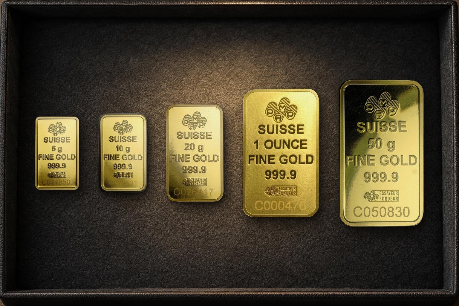 Fünf Goldbarren von PAMP nebeneinander auf dunklem Filz, von links 5, 10 und 20 Gramm, eine Unze und 50 Gramm, jeder mit den Prägungen SUISSE, FINE GOLD 999.9 und einer Nummer