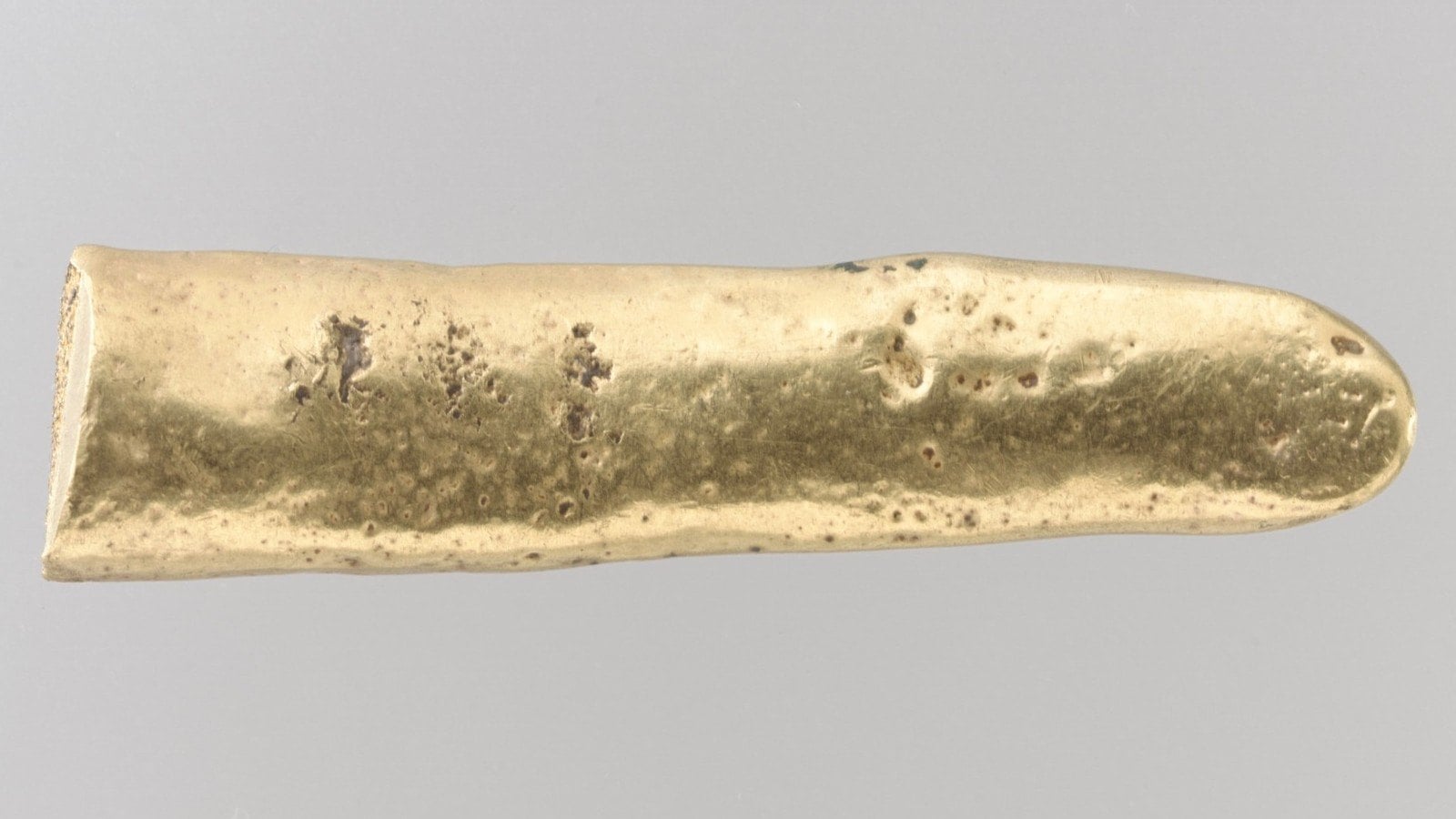 Längliches Stück Gold mit unebener, leicht narbiger Oberfläche, ein Ende gerade, das andere abgerundet, auf grauem Grund