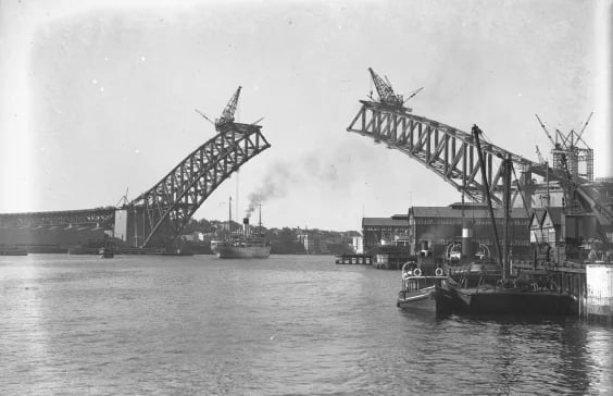 Two halves of an arch over the harbour, each with a crane at its end, a gap between them; quays on the right, water in the foreground.