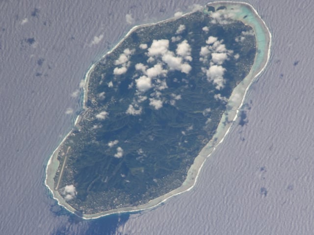 Rarotonga von der Raumstation: bewaldete Berge unter kleinen Wolken, an der Küste die Landebahn, ringsum das Riff
