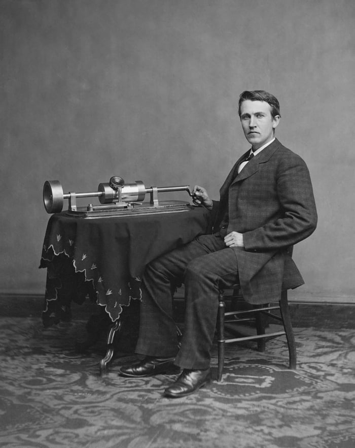 Thomas Edison sits in a suit beside a table with a dark, embroidered cloth; on it stands the phonograph with cylinder and crank, his hand rests on the crank.